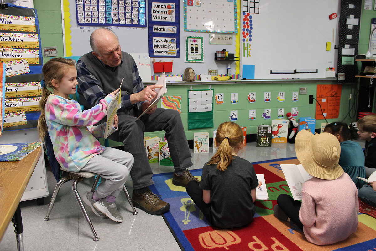 Volunteer Casey Willson sits in a chair and helps a kindergartener sitting next to him read a sentence 